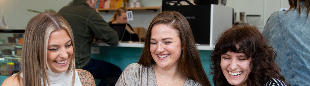 Three women sitting together at a coffee shop looking at a computer and smiling