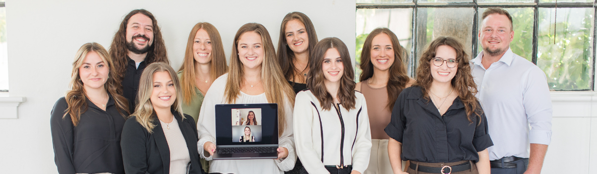 A group of people in professional clothing at a business photoshoot with two guys and eight women and one woman holding a computer with two women on zoom