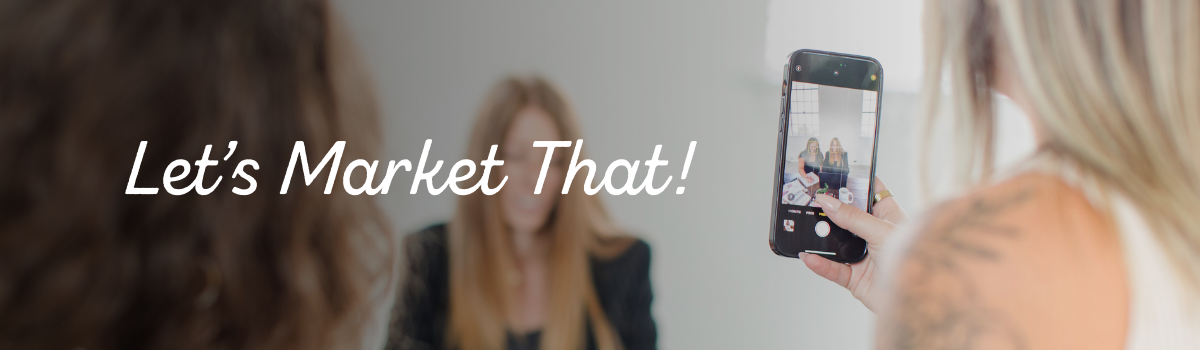 Two women in black shirts with one sitting at a desk with a computer in front of her and one on the other side holding a phone and both looking at the phone and smiling looking at a lead generation ad campaign